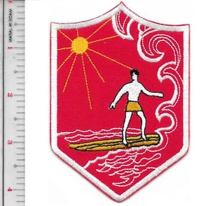 May include: A shield-shaped embroidered patch with a red background featuring a yellow sun, a white figure on a yellow surfboard, and white wave outlines. A ruler next to the patch shows it measures approximately 3.5 inches in height.