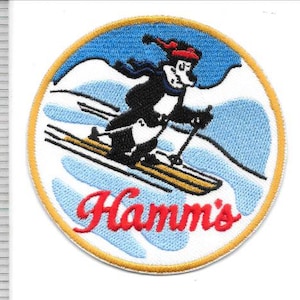 May include: Embroidered patch featuring a cartoon bear skiing down a snowy slope. The bear wears a red hat and scarf, with the word "Hamm's" in red script at the bottom. The patch has a blue and white background with a gold border.