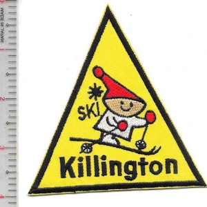 Könnte beinhalten: Gelbes dreieckiges Abzeichen mit schwarzem Rand. Es zeigt einen Skifahrer mit roter Mütze und weißem Outfit. Das Wort "SKI" steht über dem Skifahrer, und "Killington" steht darunter in schwarzen Blockbuchstaben.