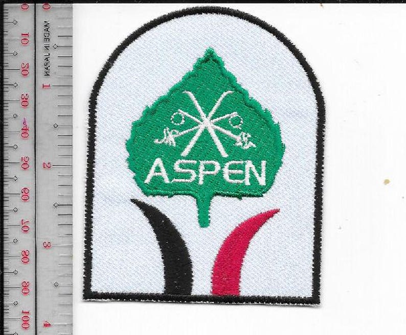 K&ouml;nnte beinhalten: Gestickter Aufn&auml;her mit wei&szlig;em Hintergrund und schwarzem Rand. Der Aufn&auml;her zeigt ein gr&uuml;nes Blattdesign mit gekreuzten Skiern und dem Wort "ASPEN" in Wei&szlig;. Unter dem Blatt befinden sich schwarze und rote gebogene Formen.