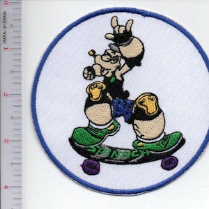 May include: Embroidered patch featuring a cartoon character skateboarding. The character is wearing green shoes, blue shorts, and yellow knee pads. The skateboard has purple wheels and the text "SPNDCH". The patch has a white background with a blue border.