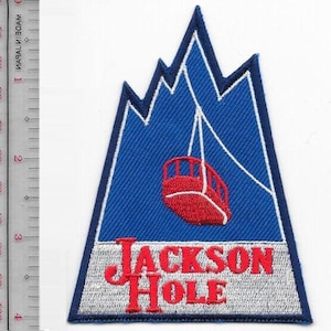 Puede incluir: Parche bordado con una cordillera azul, un teleférico rojo y las palabras "JACKSON HOLE" en rojo. El parche tiene un borde blanco y tiene forma de montaña.