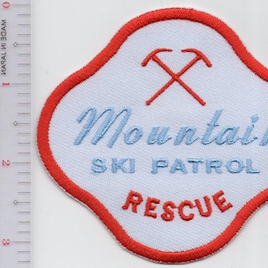 May include: A white embroidered patch with a red border, featuring two crossed red ice axes above the blue text Mountain SKI PATROL and the red text RESCUE. A clear plastic ruler is visible on the left, showing the patch is 3 inches tall.