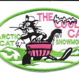 May include: Embroidered patch featuring a cartoon cat riding a snowmobile. The patch has a white background with green and pink accents. Text includes "THE COOL CAT SNOWMOBILES" and "ARCTIC CAT". The patch is approximately 3 inches wide.