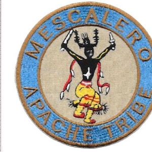 Può includere: Una toppa ricamata circolare con una figura centrale in nero, giallo e rosso, circondata da un bordo azzurro con il testo MESCALERO APACHE TRIBE. Un righello metallico mostra che la toppa ha un diametro di circa 9 cm.
