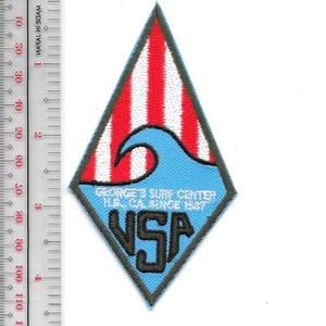 May include: A diamond-shaped patch with a blue, red, and white design. The top section features red and white stripes, while the bottom has a blue wave design and the text "GEORGE'S SURF CENTER H.B. CA. SINCE 1967" and "VSA".