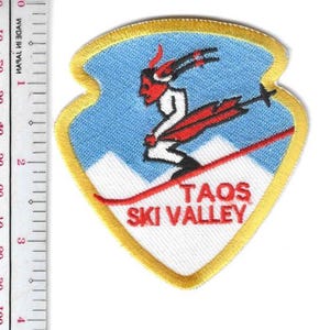 Może przedstawiać: Haftowana naszywka w kształcie tarczy, przedstawiająca narciarza z czerwoną twarzą i rogami, zjeżdżającego po czerwonym stoku. Tło jest niebieskie z białymi górami. Tekst brzmi "TAOS SKI VALLEY" na czerwono.