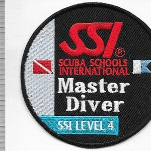 Può includere: Una toppa rotonda ricamata in bianco e nero con la scritta "SSI Scuba Schools International Master Diver Level 4". Il logo SSI è rosso. Un righello è sullo sfondo.