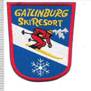 Puede incluir: Parche bordado con el texto "GATLINBURG SKI RESORT" en amarillo sobre un fondo azul y blanco. Un esquiador en rojo desciende por una pendiente amarilla. Un copo de nieve blanco está en la parte inferior. El parche tiene un borde rojo.