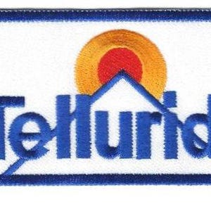Puede incluir: Parche bordado con fondo blanco y borde azul. El parche presenta la palabra "Telluride" en azul, con un diseño estilizado de montaña y sol sobre el texto. El sol es naranja y rojo.