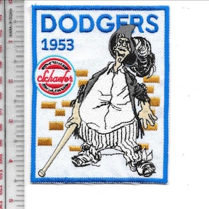 Puede incluir: Parche bordado con el texto "DODGERS 1953" en azul, y un personaje de dibujos animados sosteniendo un bate de béisbol. El parche tiene un borde azul e incluye el texto "cchaster" en un emblema circular. El personaje lleva un sombrero y pantalones a rayas.