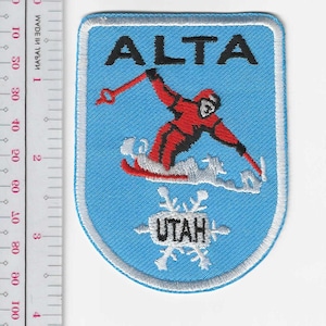 May include: Embroidered patch with a light blue background and white border. The patch features the word "ALTA" in black at the top and a skier in red and black. The word "UTAH" is at the bottom, with snowflakes.