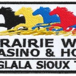 May include: A rectangular embroidered patch featuring four running horses in black, yellow, blue, and red above the text PRAIRIE WIND CASINO & HOTEL OGLALA SIOUX TRIBE. A metal ruler shows the patch is approximately 3 inches wide and 2 inches tall.