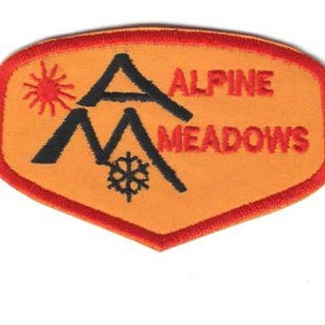 Puede incluir: Parche bordado naranja y rojo con las palabras "Alpine Meadows". El parche presenta un sol, dos picos de montaña negros y un copo de nieve. El parche tiene un borde rojo.