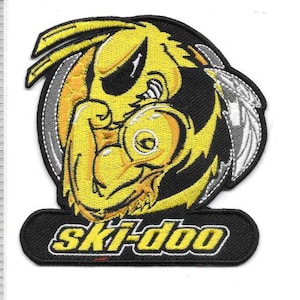 May include: Embroidered patch featuring a stylized yellow and black cartoon character flexing a bicep. The character is surrounded by a black circle with a white and silver feather design. The text "ski-doo" is at the bottom.