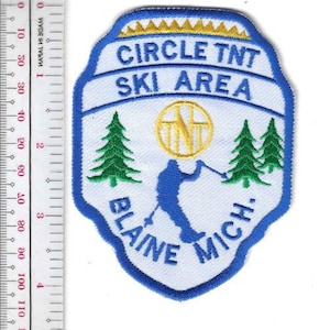 May include: A white and blue embroidered patch with the text "CIRCLE TNT SKI AREA" and "BLAINE MICH." features a skier and two green pine trees. A gold emblem is in the center. The patch is approximately 3 inches wide.