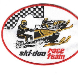 May include: Oval patch featuring two snowmobiles racing, with a checkered flag in the background. The patch has a red border and the words "ski-doo race team" in black and red. The snowmobiles are black and gold.