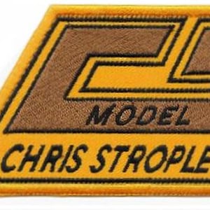 Puede incluir: Parche bordado con un diseño marrón y amarillo. El parche presenta el texto "MODEL" y "CHRIS STROPLE" en letras negras. El parche tiene una forma geométrica con un diseño "CL" marrón.