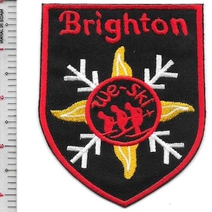 Puede incluir: Parche bordado con la palabra "Brighton" en rojo. El parche tiene forma de escudo negro con borde rojo. Un diseño de copo de nieve amarillo y blanco rodea un círculo rojo con esquiadores y el texto "we-ski".