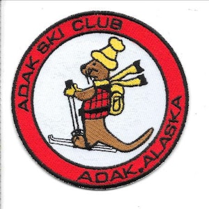 May include: Embroidered patch with a red border and the text "ADAK SKI CLUB ADAK, ALASKA." The patch features a cartoon otter wearing a yellow hat, scarf, and backpack, skiing with poles. The patch is approximately 3 inches in diameter.