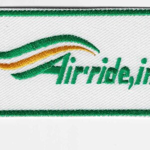 May include: Embroidered patch with a white background, green border, and green text that reads "Air'ride, inc." The patch features a design of green and gold curved lines above the text. The patch is rectangular.