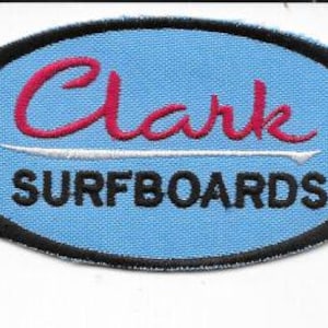 May include: An oval patch with a light blue background and black border. The word "Clark" is embroidered in red cursive above a white surfboard graphic. Below the graphic, the word "SURFBOARDS" is embroidered in black.