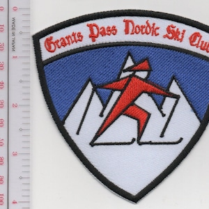 Puede incluir: Un parche bordado en forma de escudo con un cielo azul, montañas blancas y una figura de esquiador roja. El texto Grants Pass Nordic Ski Club está bordado en rojo. Una regla muestra que el parche mide unos 9 cm de alto y 9 cm de ancho.