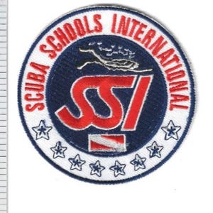 Könnte beinhalten: Runder, gestickter Aufnäher mit dem Text "SCUBA SCHOOLS INTERNATIONAL" in Rot und Weiß auf marineblauem Hintergrund. In der Mitte befinden sich die Buchstaben "SSI" in Rot, mit einer Tauchergrafik darüber. Der Aufnäher ist von weißen Sternen umrandet.