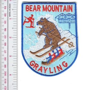 May include: Embroidered patch featuring a brown bear skiing down a snowy mountain. The patch has a light blue background with the words "BEAR MOUNTAIN" and "GRAYLING" in red and white. A skier and a ski lift are also depicted.