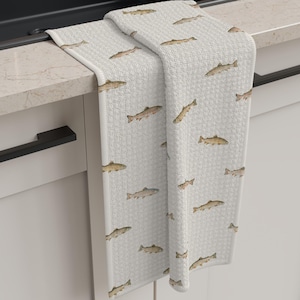 May include: White kitchen towel with a repeating pattern of brown fish. The towel is folded over a countertop next to a black sink. The towel has a textured waffle weave.