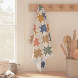 May include: A rectangular kitchen towel featuring a repeating star pattern in muted shades of blue, green, yellow, and coral on a white background. The fabric has a visible grid-like texture and is draped over a wooden wall-mounted rack.