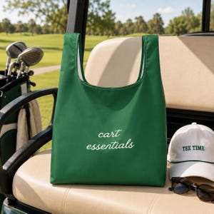 Pode incluir: Uma sacola verde com as palavras "cart essentials" em escrita branca. Um boné de beisebol branco com "TEE TIME" bordado em verde e óculos de sol repousam sobre um assento de carrinho de golfe. Os tacos de golfe são visíveis ao fundo.