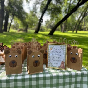 May include: A collection of brown paper gift bags decorated with bear faces featuring googly eyes and dark brown snouts, accompanied by a gold-framed sign that reads: Thank you beary much for celebrating with us! Please take one! with a bear illustration.