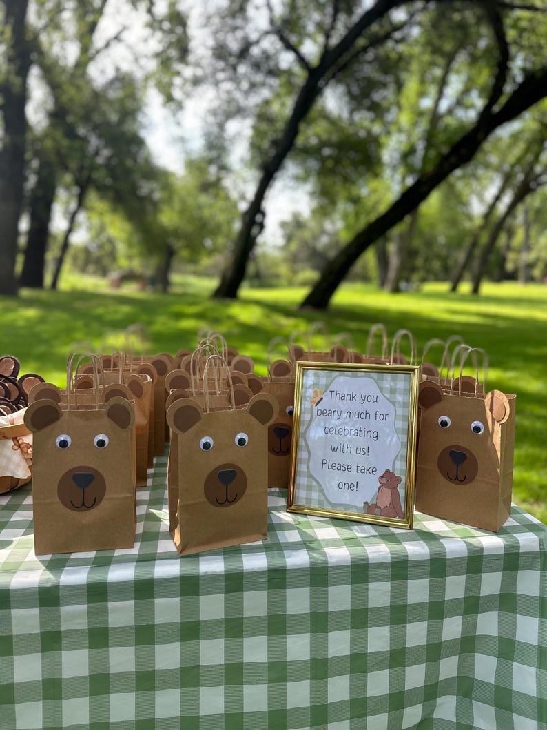 May include: A collection of brown paper gift bags decorated with bear faces featuring googly eyes and dark brown snouts, accompanied by a gold-framed sign that reads: Thank you beary much for celebrating with us! Please take one! with a bear illustration.