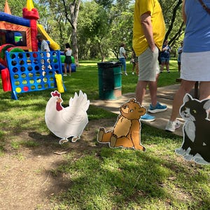 May include: Three flat, cutout-style yard signs featuring illustrations of a white chicken, a brown bear sitting down, and a black and white cat, positioned on a grassy lawn near a large blue inflatable game and a green trash barrel.