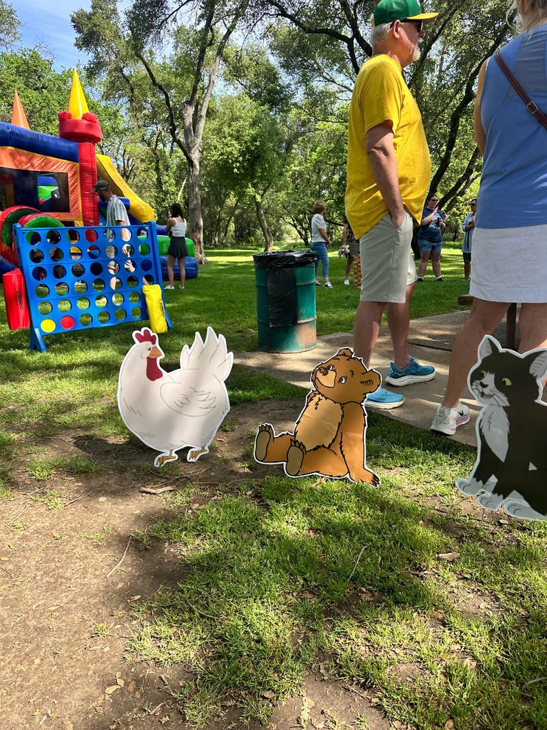 May include: Three flat, cutout-style yard signs featuring illustrations of a white chicken, a brown bear sitting down, and a black and white cat, positioned on a grassy lawn near a large blue inflatable game and a green trash barrel.