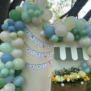 May include: A birthday display featuring a large arch of sage green, light blue, and white balloons attached to a wooden backdrop. A banner reads HAPPY BIRTHDAY in blue letters. A wooden planter box filled with white and yellow flowers sits below.
