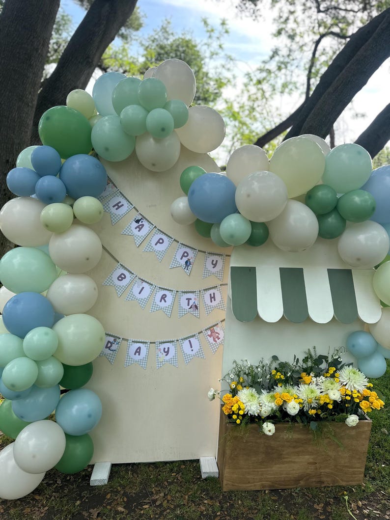 May include: A birthday display featuring a large arch of sage green, light blue, and white balloons attached to a wooden backdrop. A banner reads HAPPY BIRTHDAY in blue letters. A wooden planter box filled with white and yellow flowers sits below.
