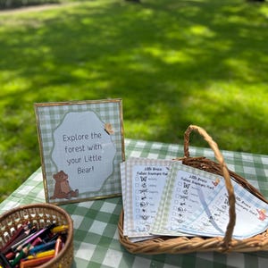 May include: A gold-framed sign with a green gingham border reads Explore the forest with your Little Bear! next to a wicker basket containing printed nature scavenger hunt checklists and a small basket filled with coloured pencils.