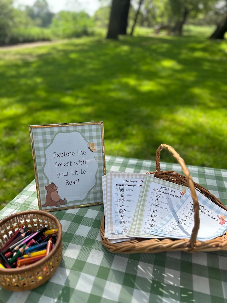 May include: A gold-framed sign with a green gingham border reads Explore the forest with your Little Bear! next to a wicker basket containing printed nature scavenger hunt checklists and a small basket filled with coloured pencils.
