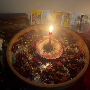 K&ouml;nnte beinhalten: Eine brennende Kerze in einer Muschelschale, umgeben von getrockneten Rosenbl&auml;ttern und Kr&auml;utern in einer Holzschale. Tarotkarten sind im Hintergrund angeordnet, zusammen mit einer roten Kerze in einem Glas. Mystisches Thema.