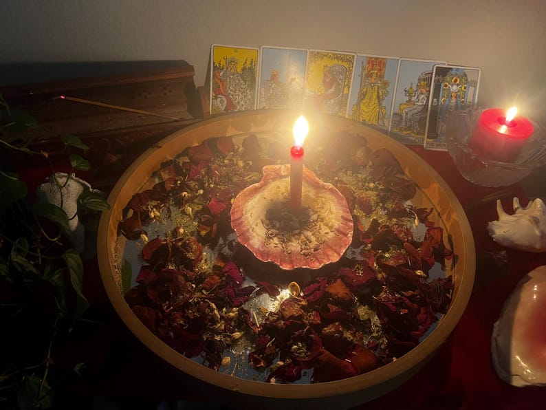K&ouml;nnte beinhalten: Eine brennende Kerze in einer Muschelschale, umgeben von getrockneten Rosenbl&auml;ttern und Kr&auml;utern in einer Holzschale. Tarotkarten sind im Hintergrund angeordnet, zusammen mit einer roten Kerze in einem Glas. Mystisches Thema.