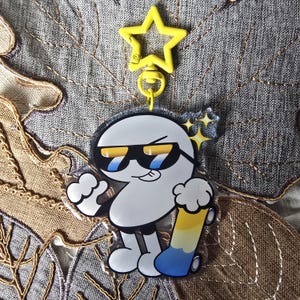 May include: A white cartoon character keychain with sunglasses and a skateboard. The keychain has a yellow star-shaped clip. The skateboard is yellow and blue. The character is smiling and has a thumbs up.