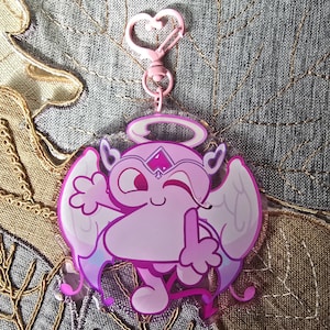 May include: A pink acrylic keychain featuring a cartoon angel with a halo, wings, and a devil tail. The angel is winking and giving a thumbs up. The keychain has a heart-shaped clasp.