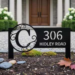 May include: A black metal address sign featuring a large circular cutout with a decorative scrollwork letter C, followed by the numbers 306 and the text HIDLEY ROAD in a stencil-style font, designed for placement in a garden or lawn area.