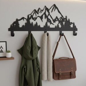 May include: Black metal wall-mounted coat rack featuring a mountain and forest silhouette design. The rack has four hooks and is holding a green jacket, a cream-colored scarf, and a brown leather satchel. A small shelf with a plant is visible.