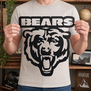 May include: A light grey short-sleeved t-shirt featuring a large black graphic of a roaring bear head centred on the chest. Above the bear, the word BEARS is printed in bold, black, capital letters within a rectangular border design.