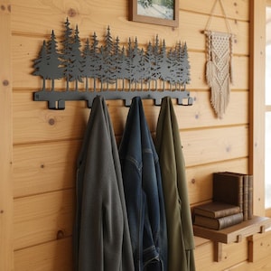 May include: A matte black metal wall-mounted coat rack featuring a laser-cut silhouette of a dense pine forest along the top edge. The rack includes five downward-facing hooks for hanging garments, currently holding three jackets in gray, blue, and olive.