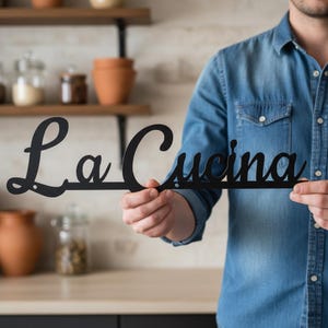 May include: A black metal wall sign featuring the word La Cucina in a cursive script font. The sign is constructed from a single piece of thin, dark material with a smooth finish and includes small circular mounting holes at the ends.