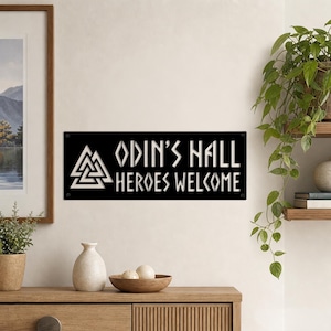 Può includere: Una targa rettangolare in metallo nero con il simbolo Valknut bianco sulla sinistra e il testo "ODIN'S HALL HEROES WELCOME" in un carattere stilizzato. La targa presenta quattro piccoli fori di montaggio circolari, uno per ogni angolo.